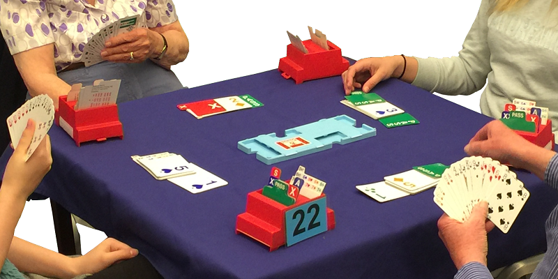 A blue, cloth covered table top with four sets of cards and bidding boxes (boxes for cards showing bids) for a brige card game. 