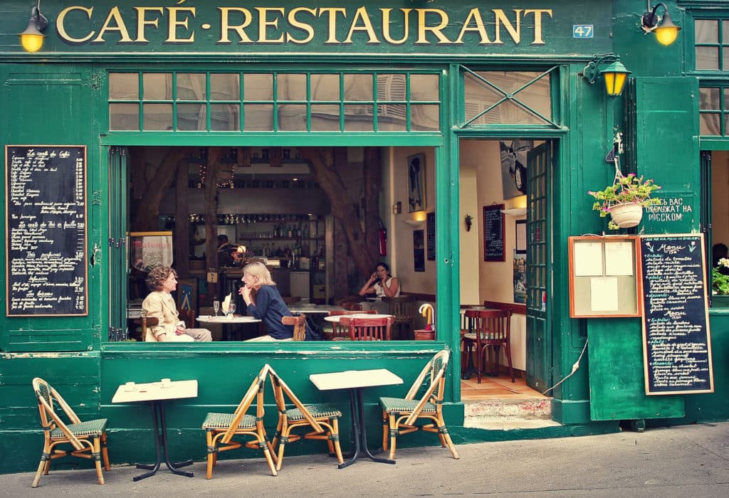 View through an open door and large window of a French cafe showing two women talking over a table.  Tables and chairs outside.  Whole shop facade coloured in the same rich green.