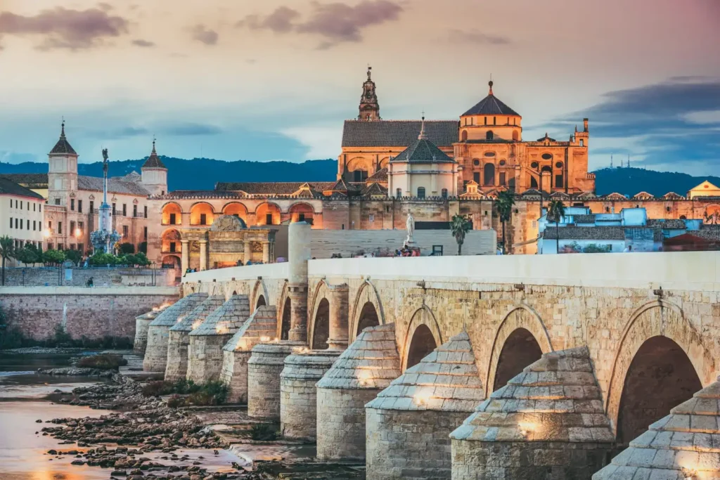 View across a wide, white stone, multi-arched bridge over a dryish river, showing a typical, large church highlighted orange by the setting sun. 