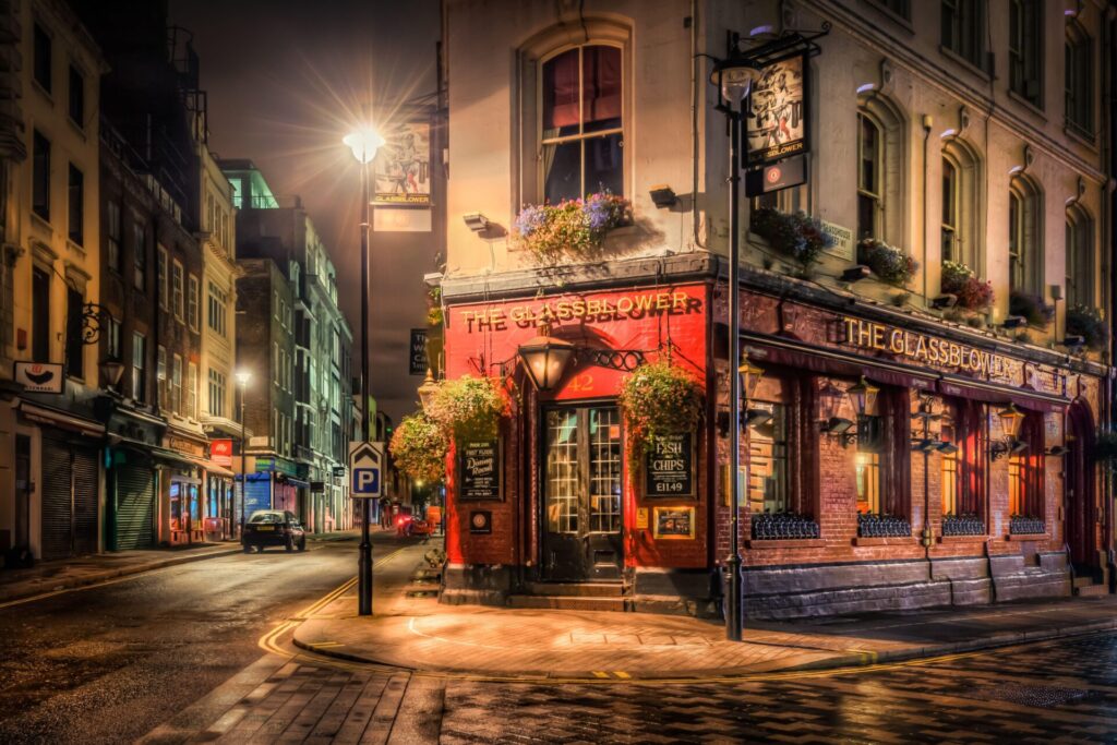 Noght scene of The Glassblower pub in Piccadilly, London. Narrow wedge shaped building. Red painted wall. Dark wooden doors with two thirds covered in small pane windows. Hangin baskets and Fish and Chips signs on the wall.