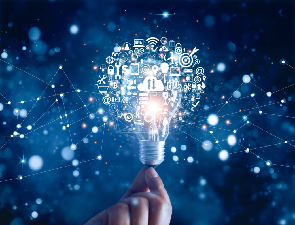 A tungsten light bulb balanced on the finger tips of a hand and swithed on dimly. Around the glass part is a circular area full of small white and clear icons of many objects, mainly related to technology, such as mobile phone, spanner, gears, wi-fi sign. All on a blue background with various white dots from small clear to large bokeh, like the sky at night.