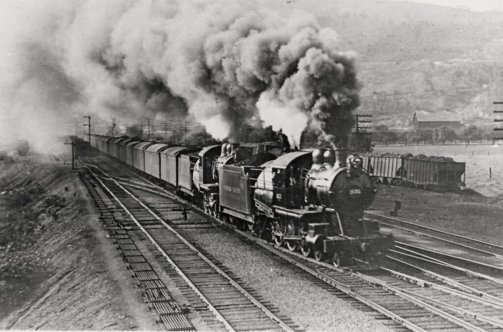 Black and white photo. Two 460 steam locomotives pulling a long goods train over points past a yard. Lots of dirty steam belching across the view.